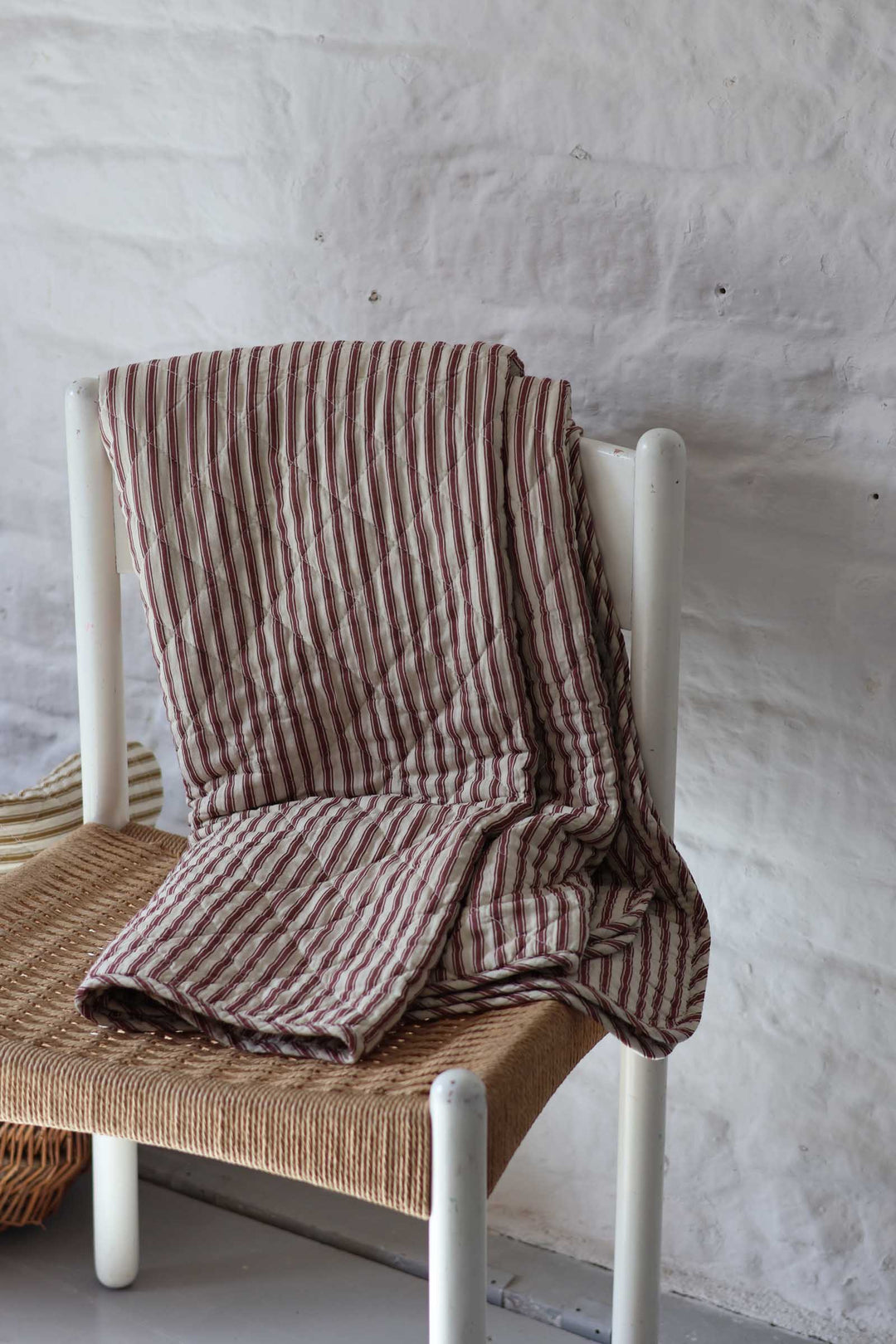 A striped quilted blanket in brown and beige is draped over a white wooden chair with a woven seat, set against a textured white wall.