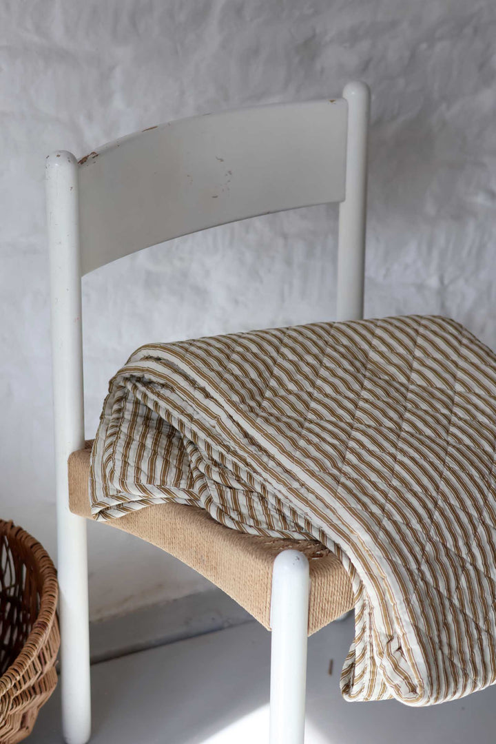 A folded striped quilted blanket in beige and white is draped over a white wooden chair in a rustic room setting.