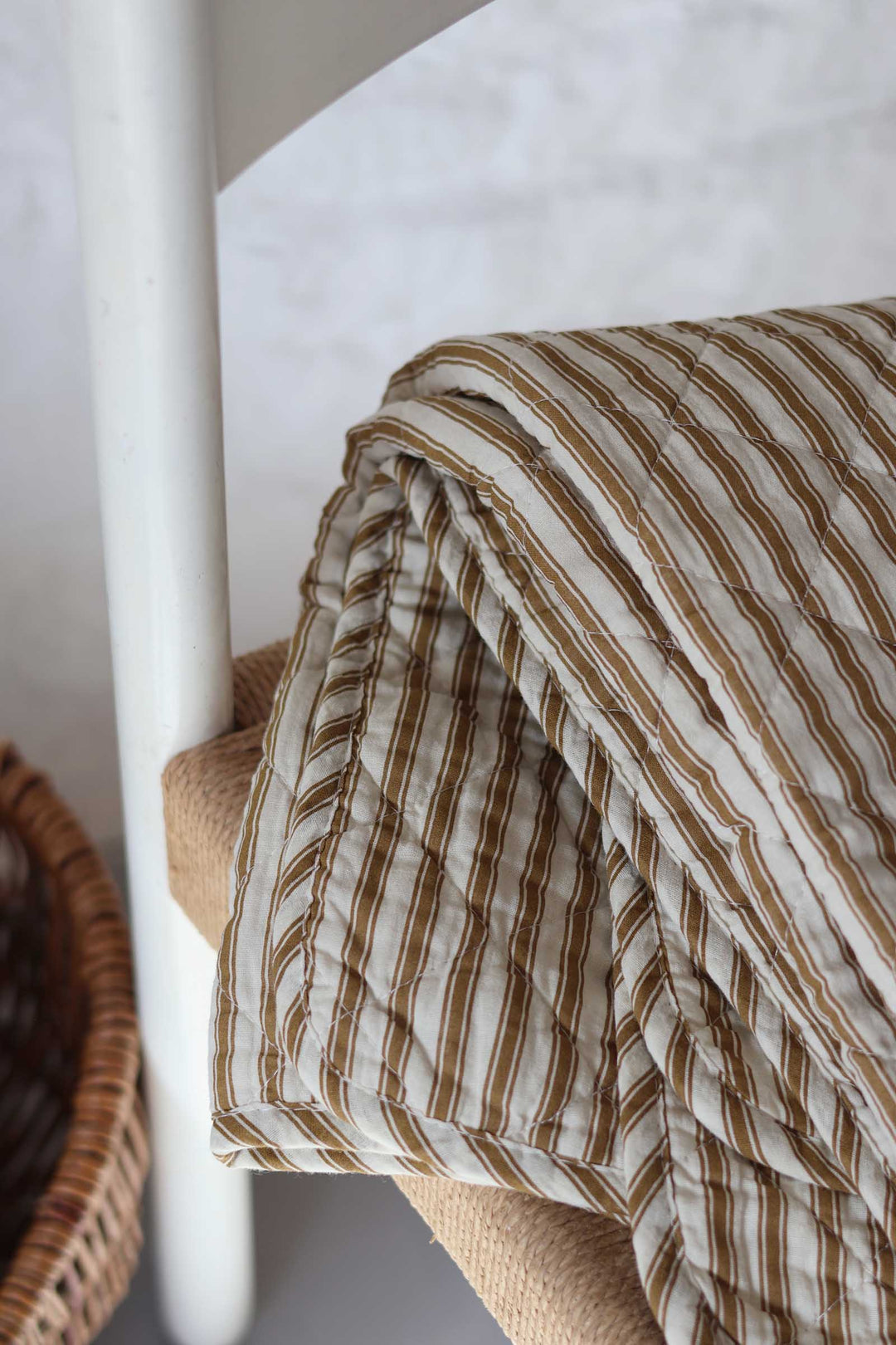 A close-up of a quilted blanket with brown and cream vertical stripes draped over a woven chair, with a white chair leg and a wicker basket visible in the background.