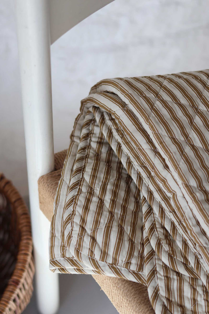 A close-up of a quilted blanket with brown and cream vertical stripes draped over a woven chair, with a white chair leg and a wicker basket visible in the background.