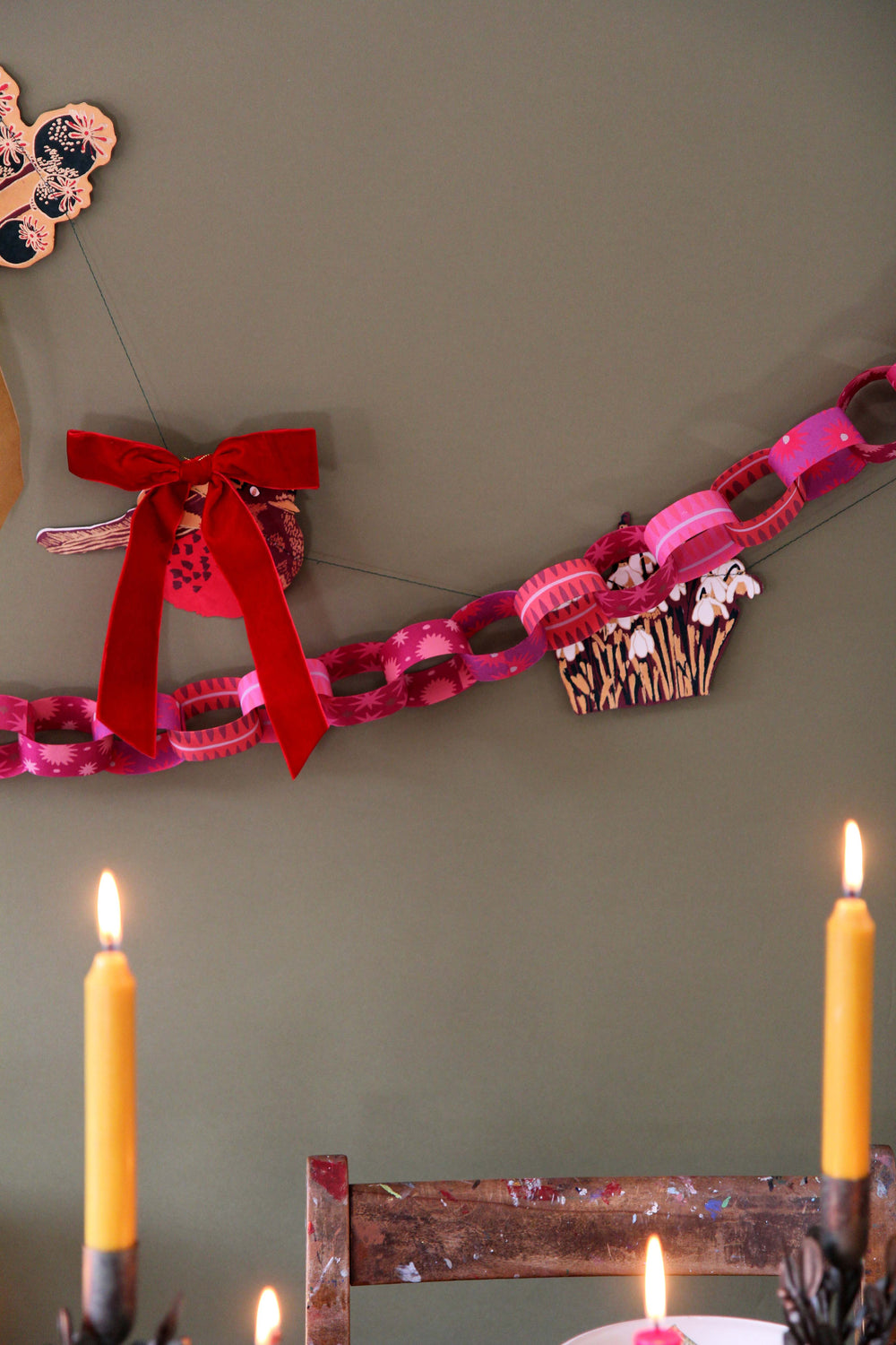 Decorative paper chain with a red bow and candles on a green wall.