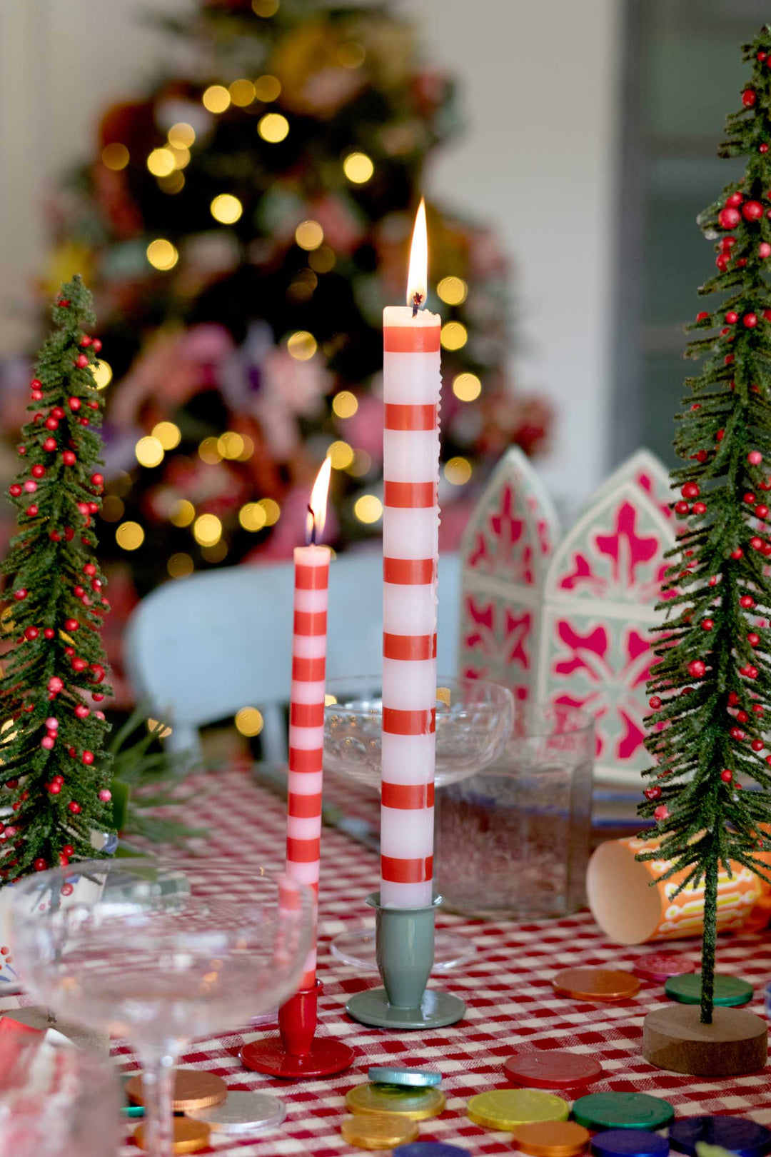 Two red and white striped Christmas taper candles in metal holders on a red-and-white checkered tablecloth, with blurred holiday decorations and a Christmas tree in the background.