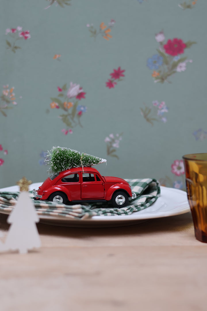 A miniature red toy Volkswagen Beetle car carrying a small green Christmas tree, styled on a white plate with a green plaid napkin on a wooden table against a floral wallpaper background.