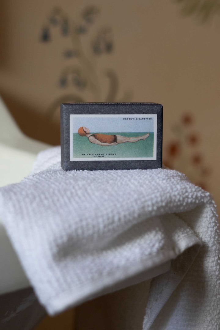 A vintage matchbox featuring a medical illustration of a drowned body, resting on a white textured towel against a warm, blurred background.