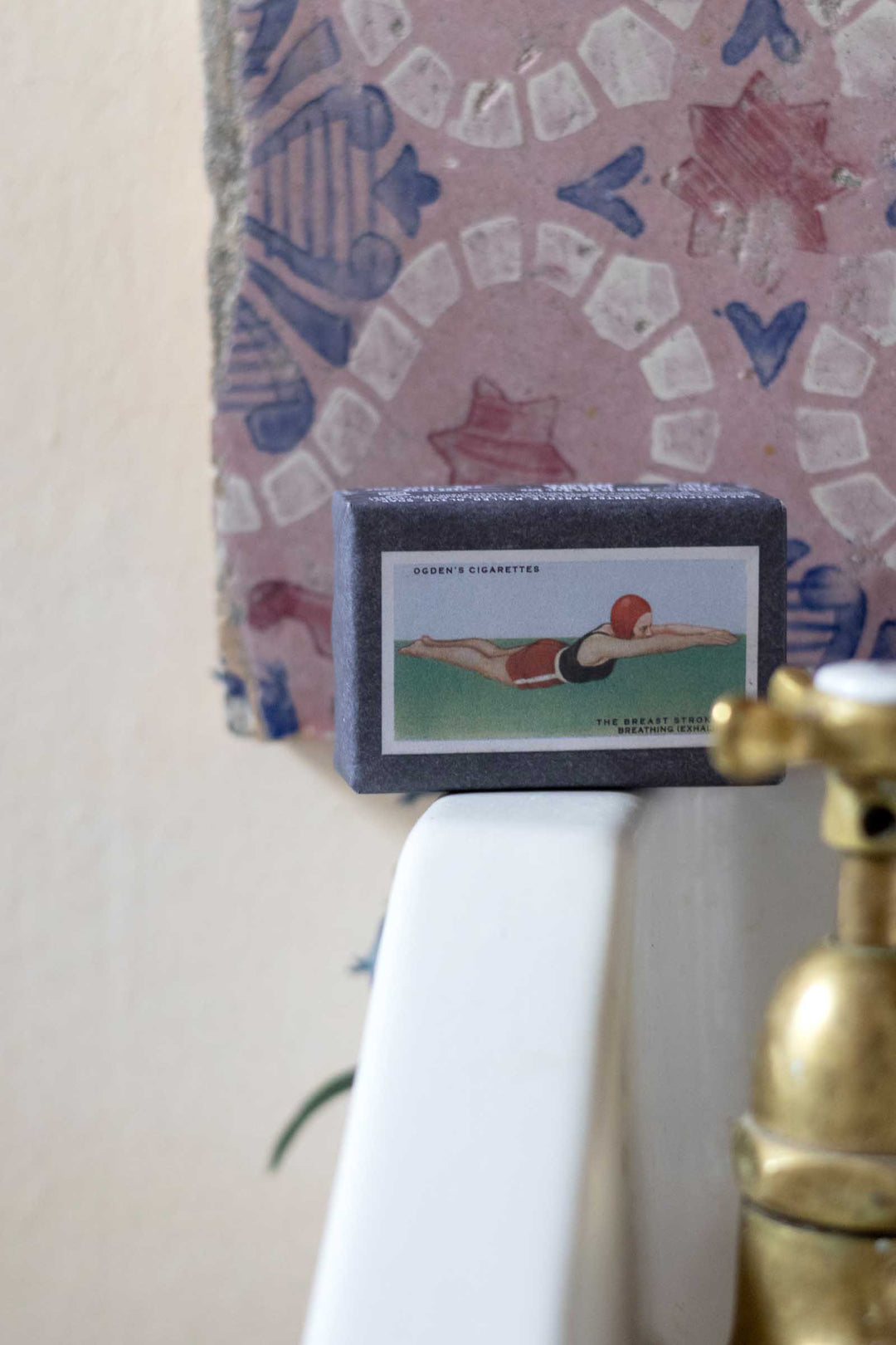 A vintage collectible matchbox featuring a 1912 Coca-Cola advertisement, displayed on a white ceramic ledge next to a brass faucet.