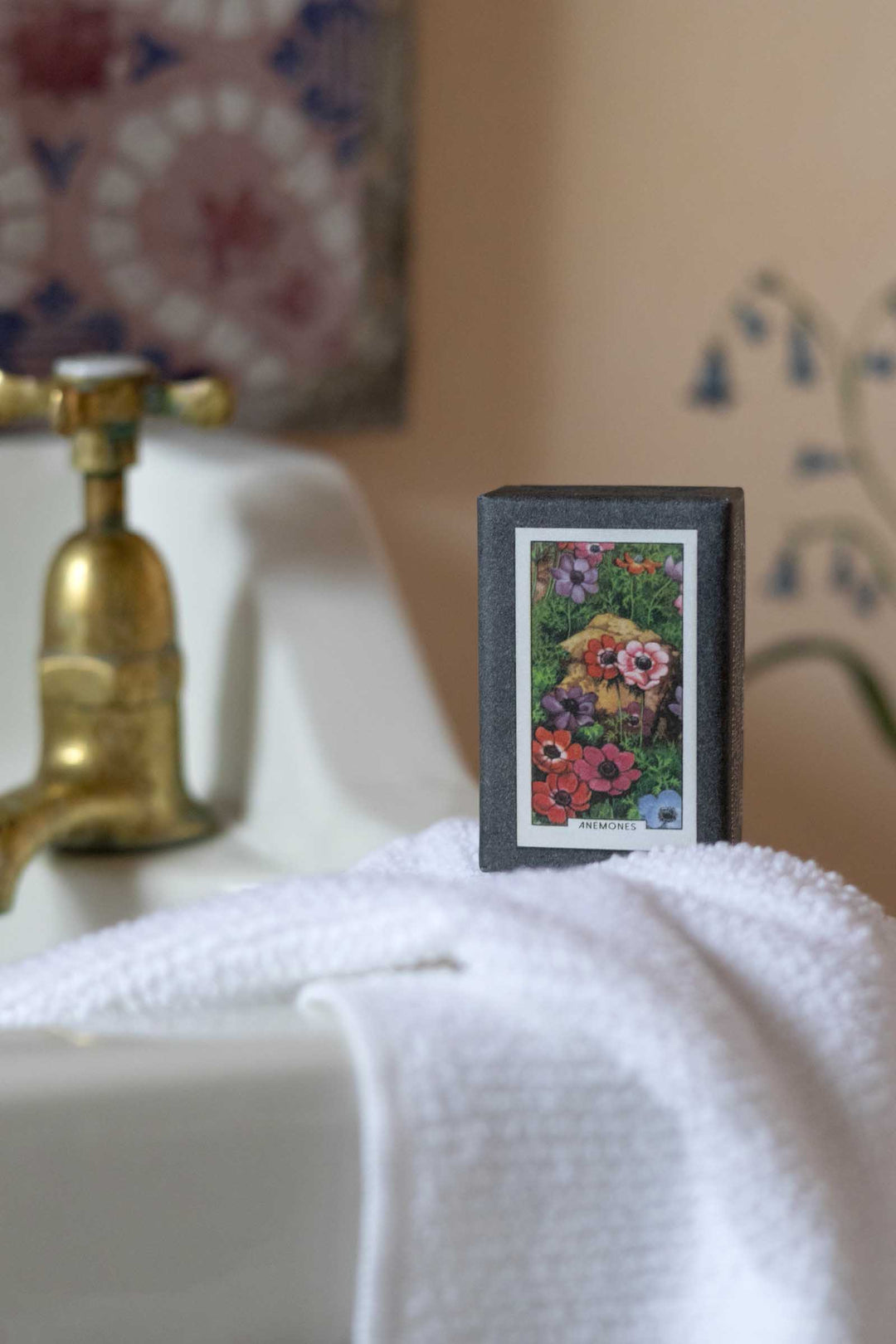 A small framed floral art print with a dark gray frame and white mat, resting on a white towel next to a brass faucet in a bathroom setting.