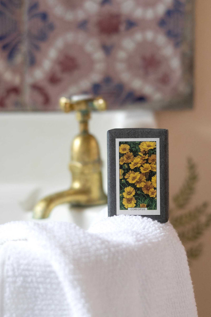 A rectangular soap bar with a dark gray border and a yellow flower print, standing on a white towel in front of a brass faucet and patterned tile background.