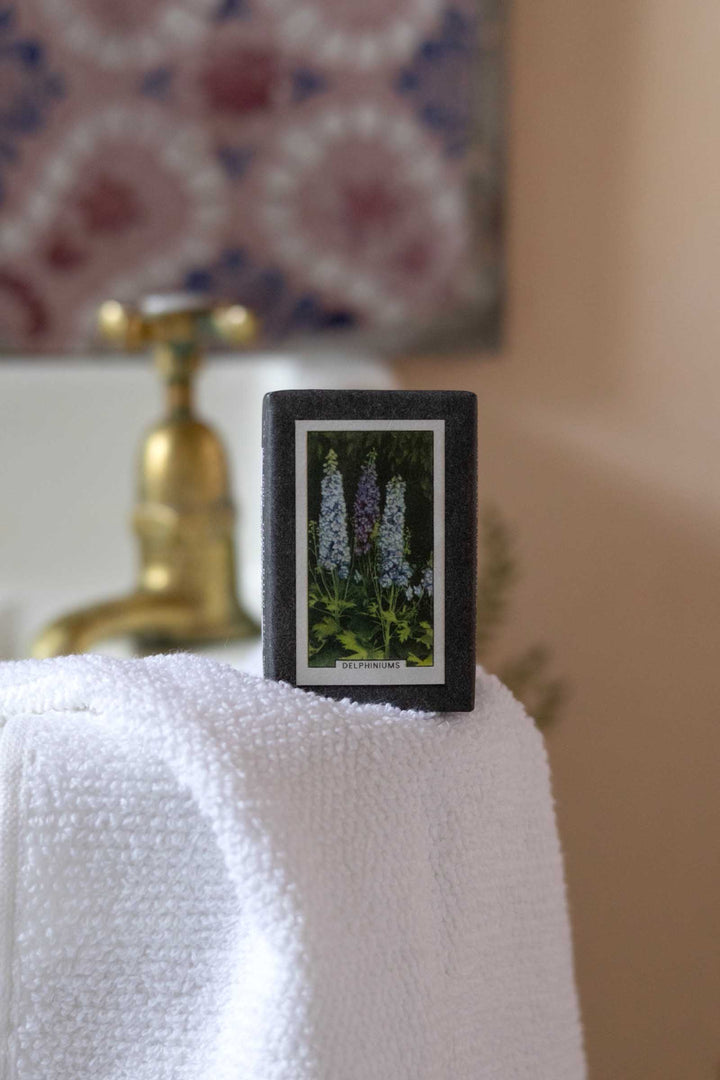 A small rectangular black-framed floral print or card standing on a white towel, with a blurred bathroom faucet and patterned wall in the background.
