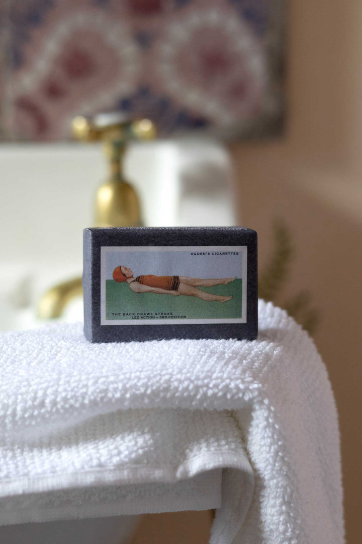 A rectangular vintage collectible card or print with a dark border, featuring an illustration of a woman in a swim cap and swimsuit on a green background, displayed on a white textured towel in a softly blurred bathroom setting.