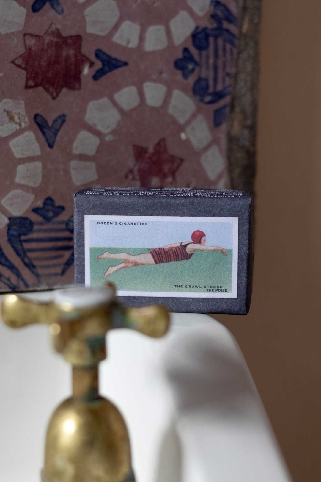A vintage cigarette pack box labeled 'Duby's Cigarettes' with an illustration of a woman in a red swimsuit, resting on a white surface in front of a patterned mosaic tile background.