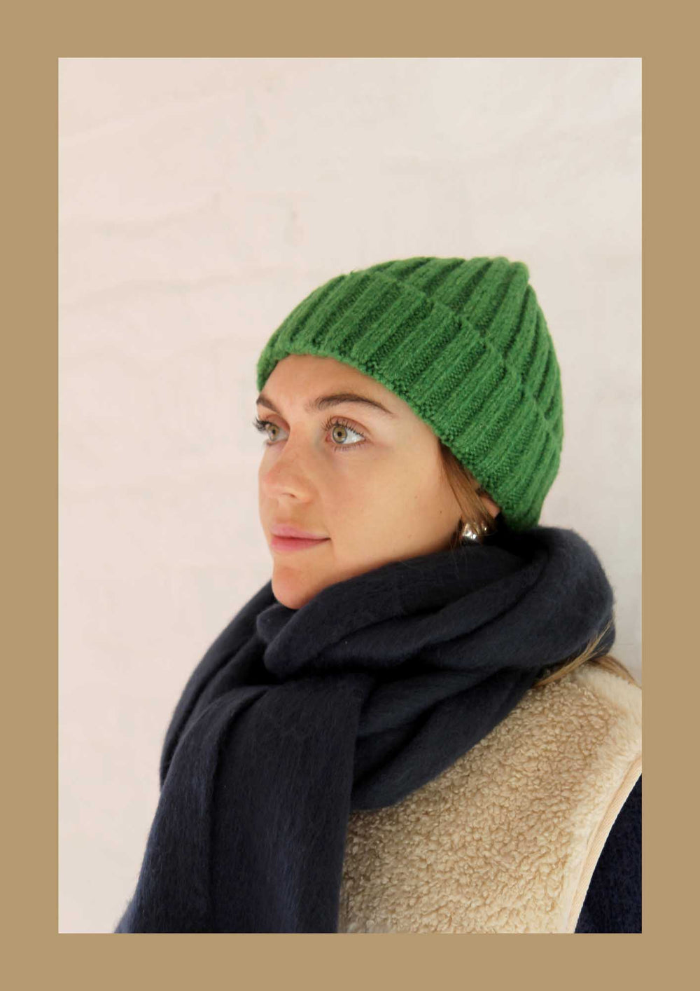 Person wearing a green knitted beanie and dark scarf against a beige background