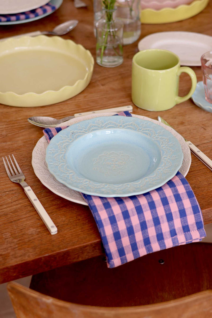 Dining table setting with a blue plate, pink and white checkered napkin, and green mug.