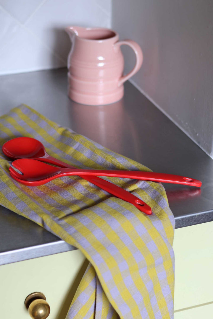 Red plastic kitchen utensils, including a spoon and spatula, resting on a yellow and white striped kitchen towel on a stainless steel countertop, with a pink ceramic pitcher in the background.