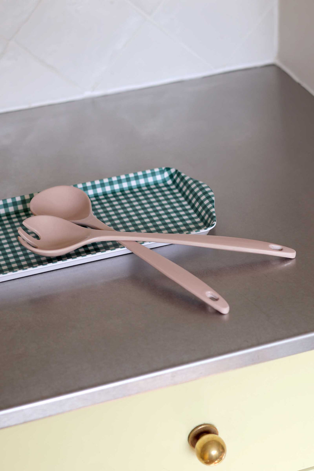 Two matte blush pink kitchen utensils, likely spoons or spatulas, resting on a green and white checkered tray on a brushed metal countertop.