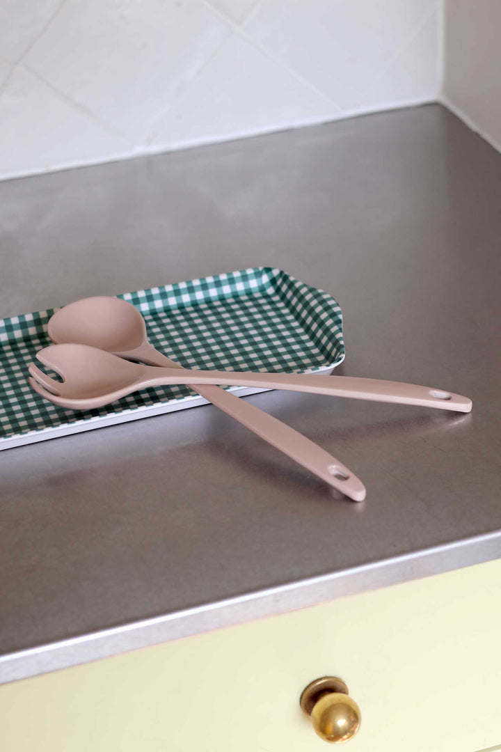 Two matte blush pink kitchen utensils, likely spoons or spatulas, resting on a green and white checkered tray on a brushed metal countertop.