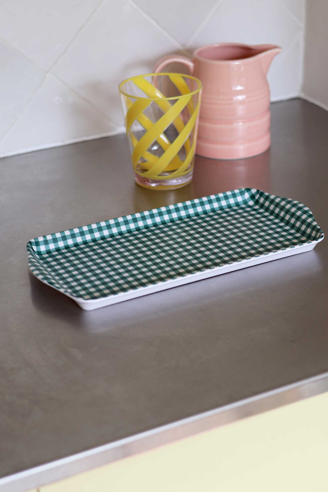A rectangular serving tray with a green and white gingham check pattern, shown on a stainless steel countertop next to a yellow patterned glass and a pink pitcher.
