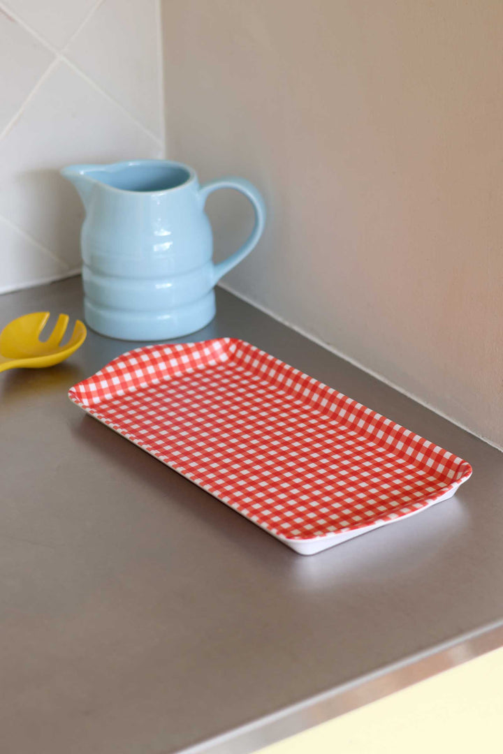 A rectangular serving tray with a red and white gingham checkered pattern, placed on a gray countertop next to a light blue ceramic pitcher and a yellow spoon.