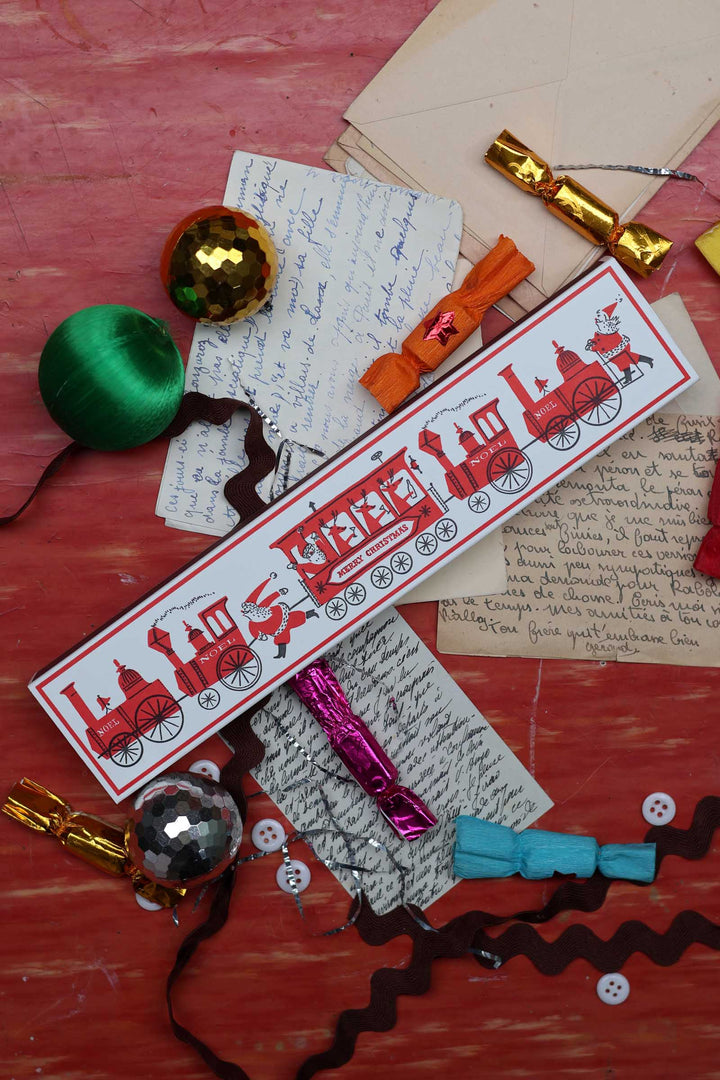 A festive Christmas-themed gift tag featuring a red train illustration on a white background with red text, styled in a flat-lay arrangement with holiday ornaments, handwritten letters, and candy on a red wooden surface.