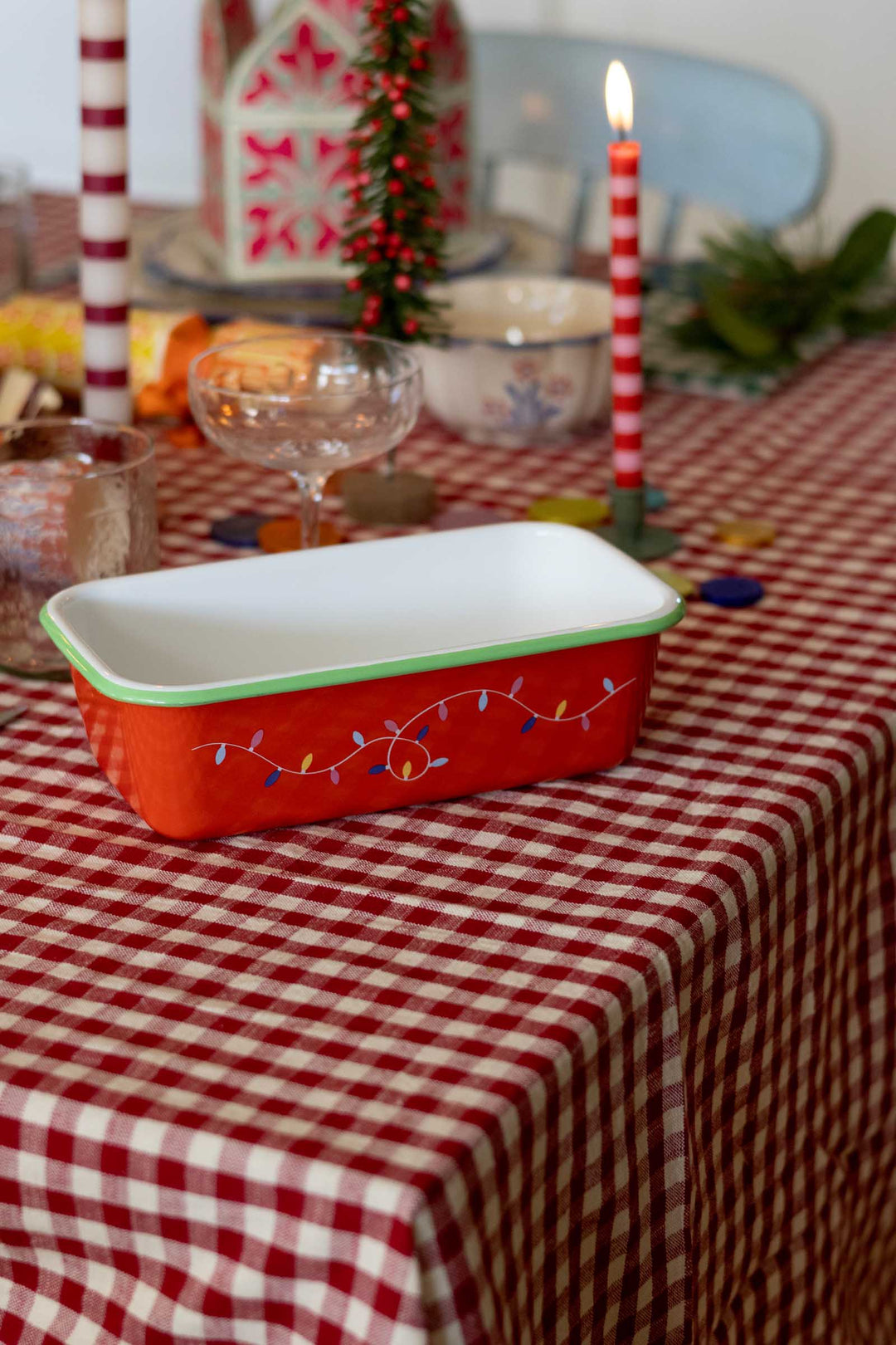 A red rectangular enamel baking dish with a white interior and a green rim, decorated with colorful Christmas string lights, set on a red and white checkered tablecloth with candles in the background.