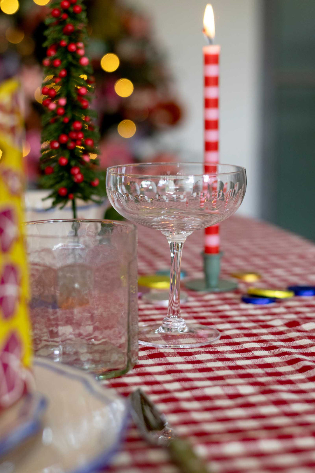 A festive holiday table setting with a red and white checkered tablecloth, a clear stemmed coupe glass, a matching textured tumbler, a lit red and white striped candle, and a small decorated Christmas tree in the background.