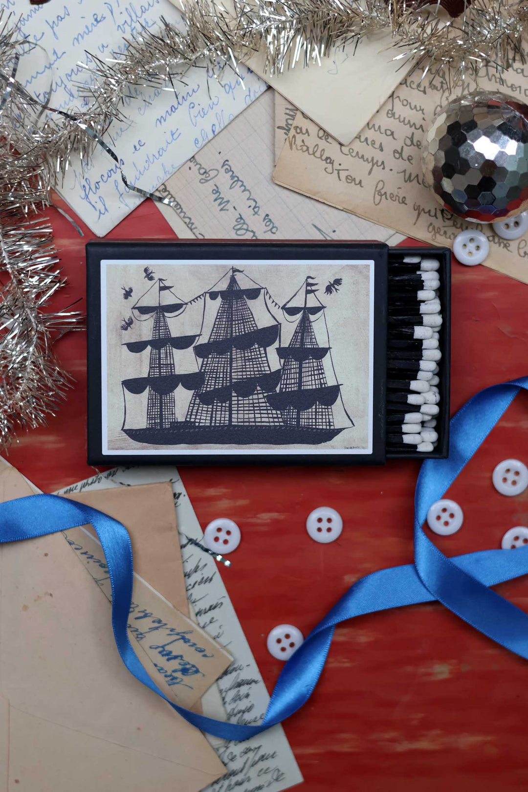 A decorative matchbox-style tin featuring a navy blue ship illustration on a cream background, styled on a red wooden surface with handwritten letters, silver tinsel, white buttons, blue ribbons, and a disco ball.