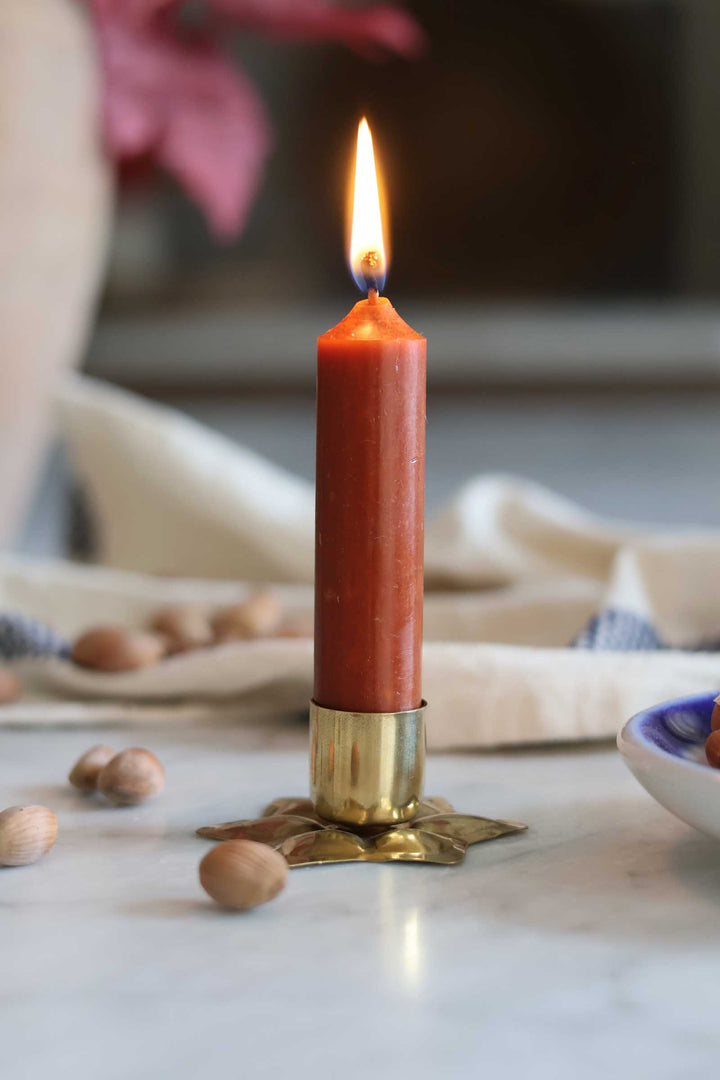 A lit orange-red taper candle in a brass star-shaped holder, set on a marble surface with scattered hazelnuts and a softly blurred background.