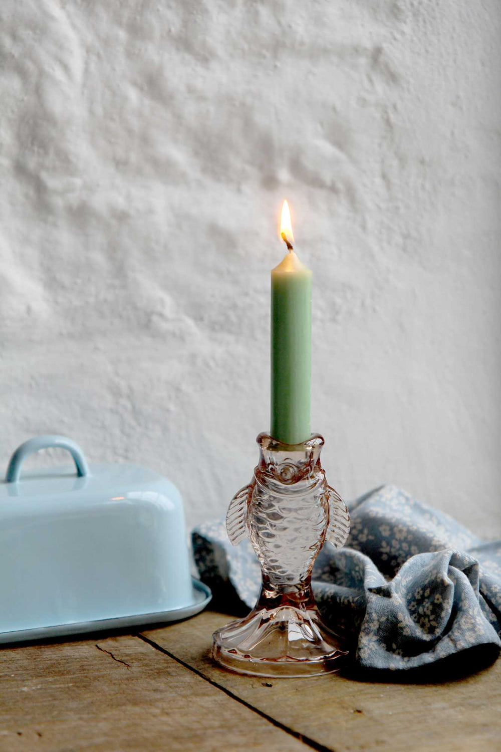 Green candle in a decorative glass holder on a wooden surface with a light blue dish and gray towel in the background.