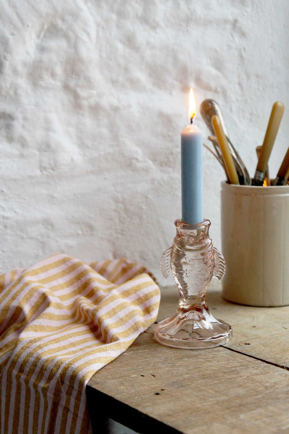 Candle in a glass holder on a wooden surface with a striped cloth and a container in the background.
