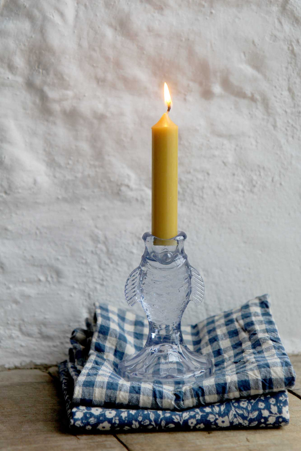 Clear glass candle holder with a lit yellow candle on a stack of blue and white checkered cloths against a textured wall.