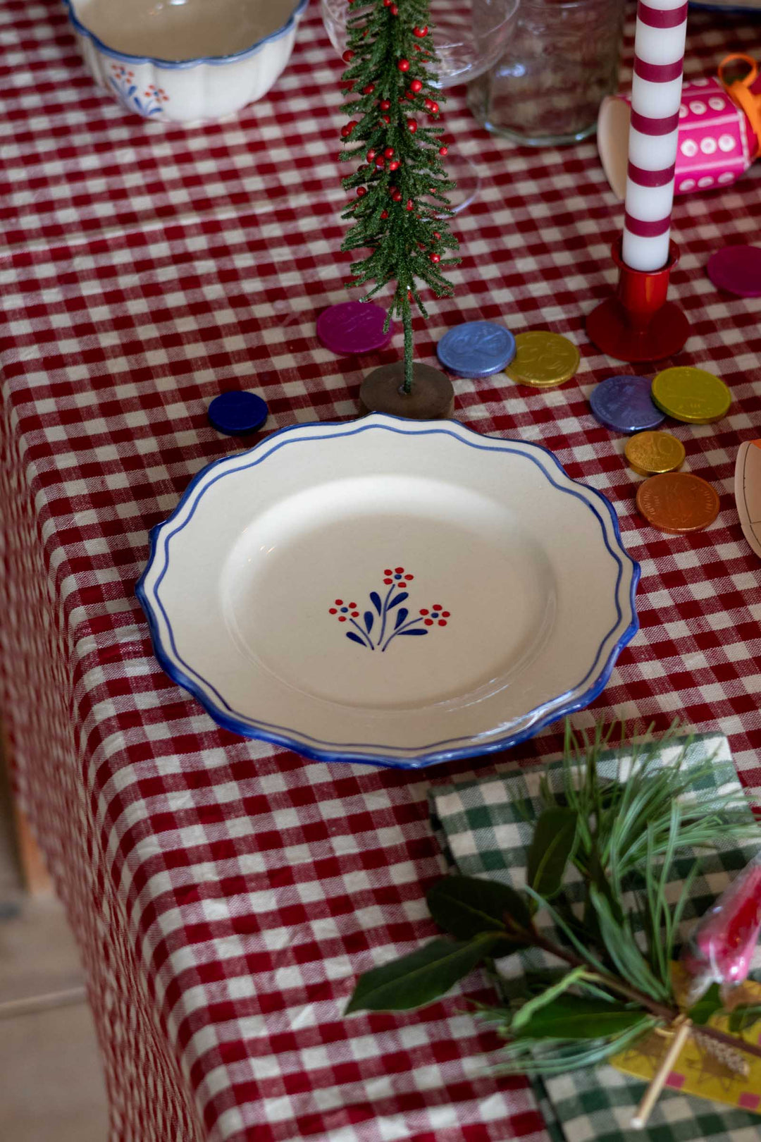 A white ceramic plate with a blue scalloped rim and a small floral motif in the center, set on a red and white checkered tablecloth alongside holiday decorations.