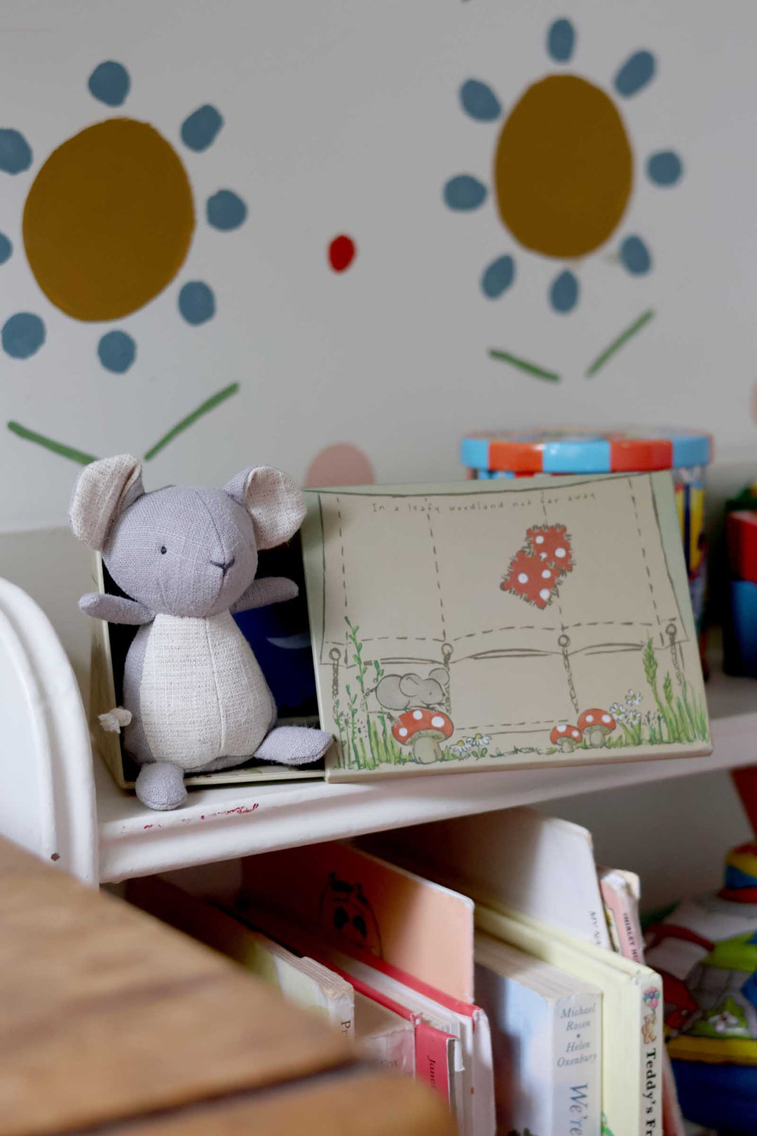 Children's room with a shelf holding books and a mouse plush toy, with a colorful wall in the background.