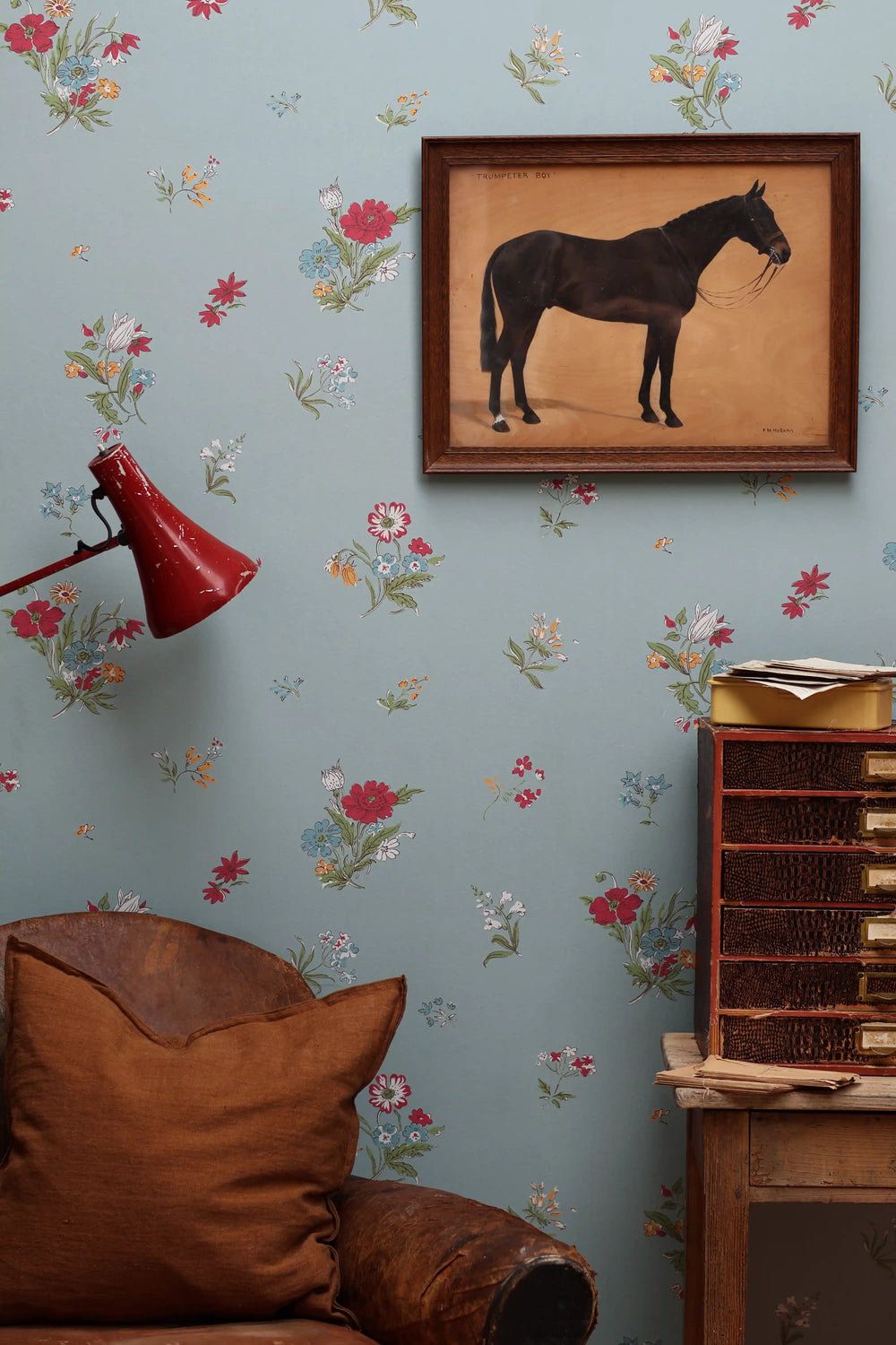 A styled interior scene featuring light blue wallpaper with a delicate multicolor floral pattern, a framed vintage painting of a black horse, a red wall lamp, and a brown leather armchair.