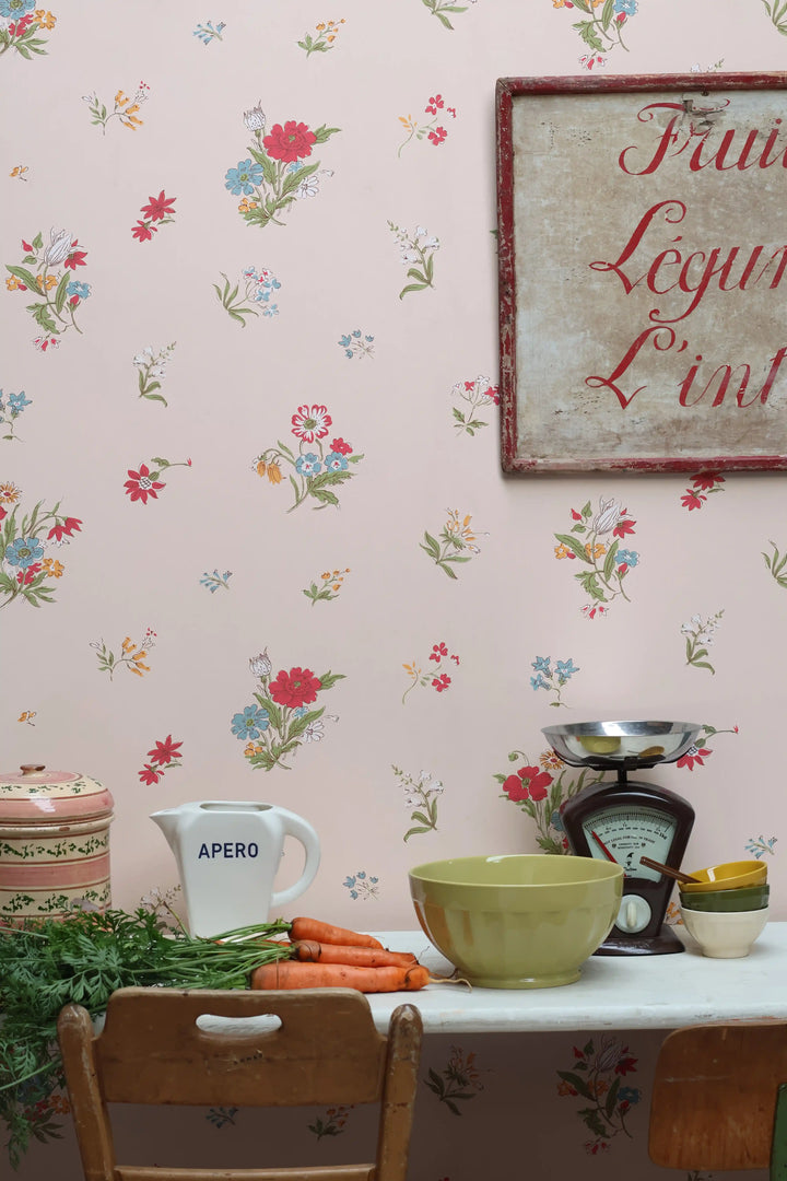 A styled kitchen or dining corner with light powder pink floral wallpaper, a vintage-style framed sign on the wall, a white table, wooden chairs, a green bowl, carrots, a white pitcher labeled APERO, and a small scale.