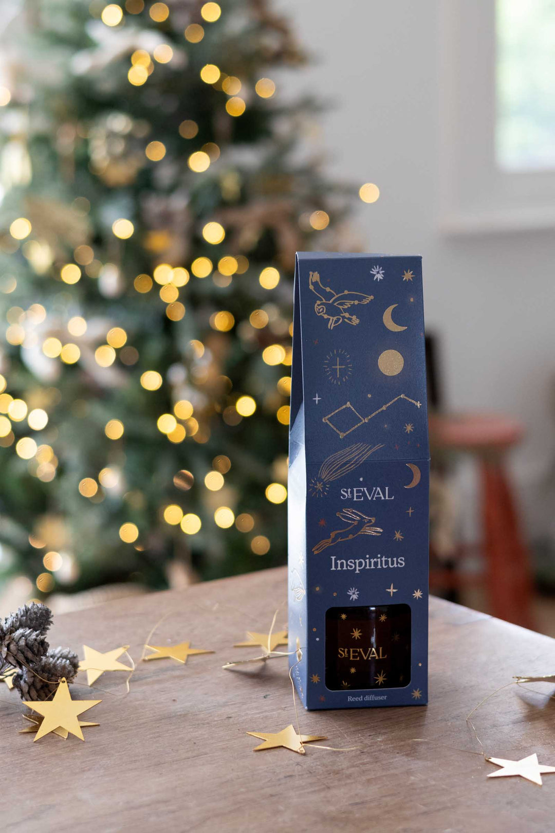 A tall dark blue SEVAL Inspiritus gift box with gold celestial illustrations stands on a wooden table in a cozy indoor setting with a blurred Christmas tree and warm lights in the background.