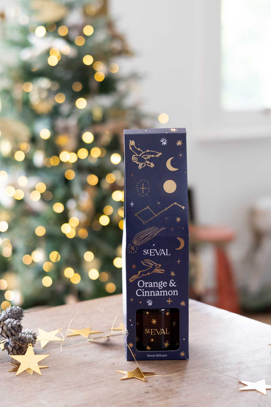 SEVAL Orange & Cinnamon scented diffuser in its dark navy blue box packaging, decorated with gold celestial illustrations, set on a wooden table with a blurred Christmas tree in the background.