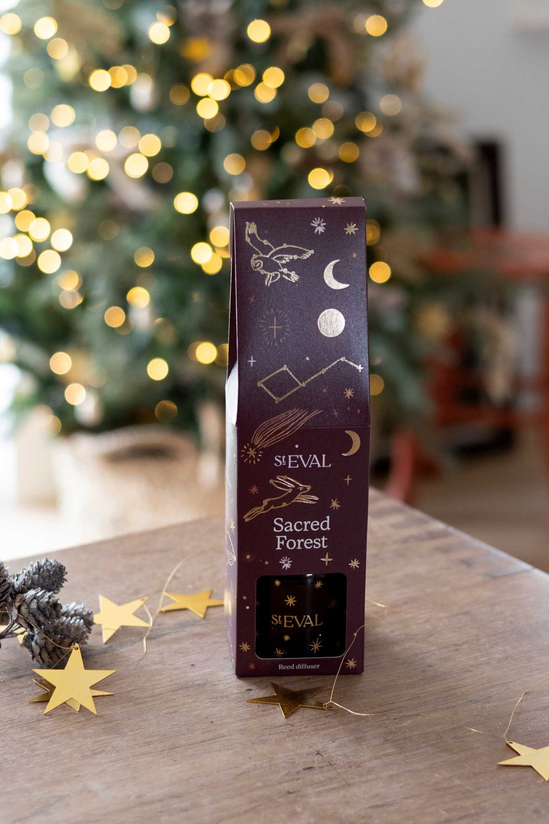 A tall, narrow SEVAL 'Sacred Forest' perfume box stands on a wooden table, decorated with gold celestial illustrations on a dark plum background. The scene is set in a cozy indoor environment with a blurred Christmas tree in the background and gold star decorations on the table.