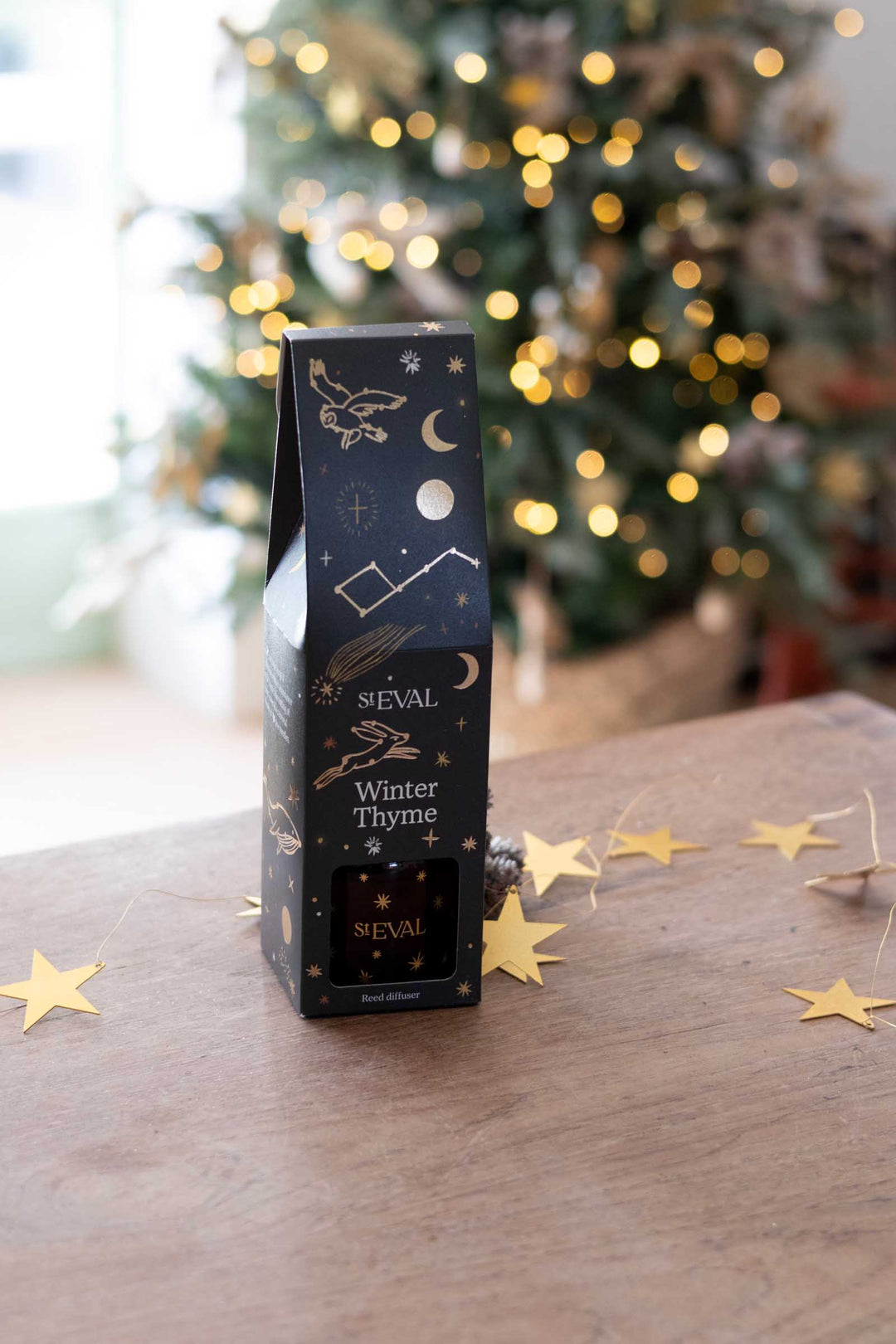 A tall black box of Séval Winter Thyme with gold celestial illustrations, standing on a wooden table with a blurred Christmas tree in the background.