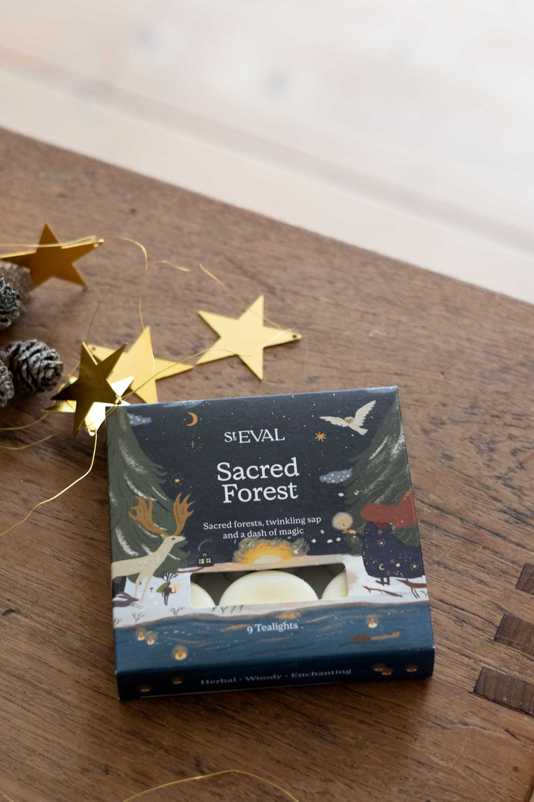 A boxed soap product labeled 'SIEVAL Sacred Forest' sits on a wooden tabletop with gold star garlands and pinecones nearby. The box is dark blue with illustrated winter forest and animal artwork, and the soap bars are visible through a window on the front.