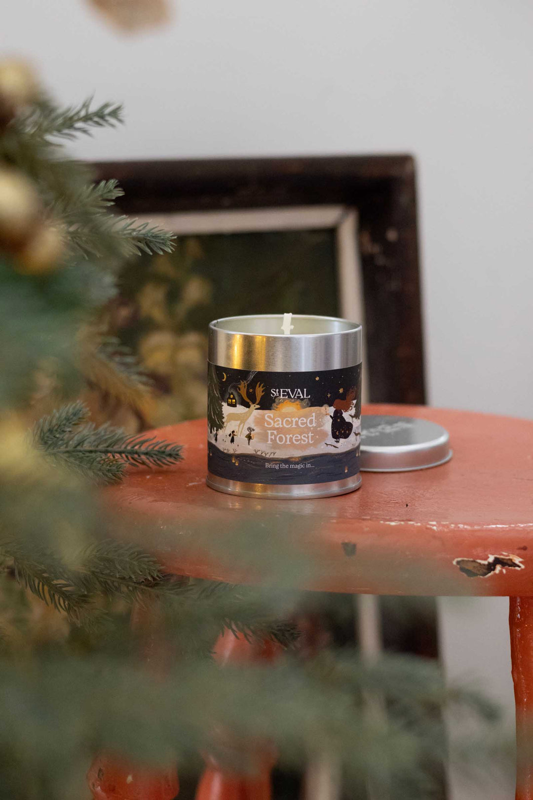A silver metal tin candle labeled 'SEVAL Sacred Forest' sits on a distressed orange-red wooden stool, with a pine tree branch in the foreground and a dark picture frame in the background.