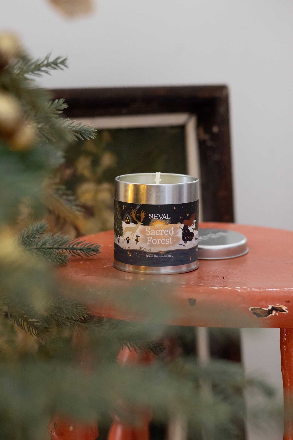 A silver metal tin candle labeled 'ST EVAL Sacred Forest' sits on a distressed orange-red wooden stool, with a pine tree branch in the foreground and a dark picture frame in the background.