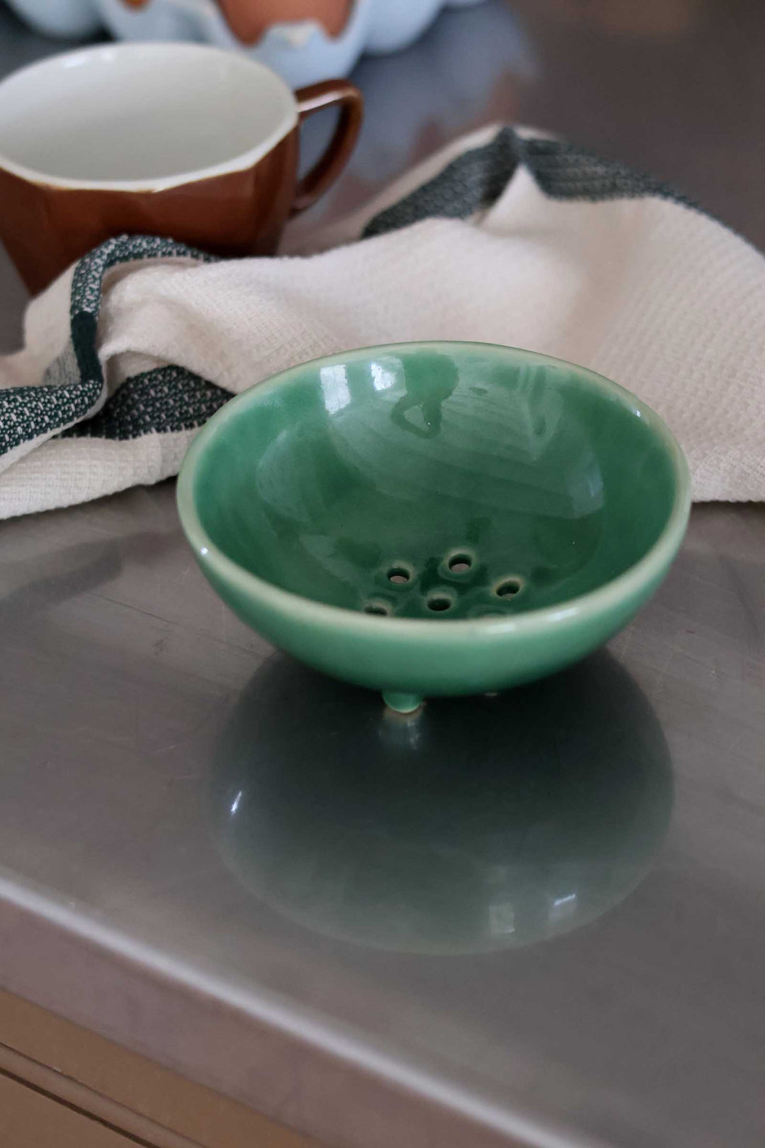 A small glossy green ceramic bowl with footed legs and small drainage holes, placed on a reflective gray tabletop with a cream cloth and a brown mug in the background.