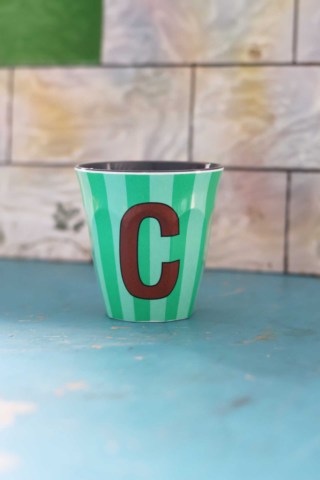 Green and white striped cup with a brown letter 'C' on a blue surface and tiled wall background