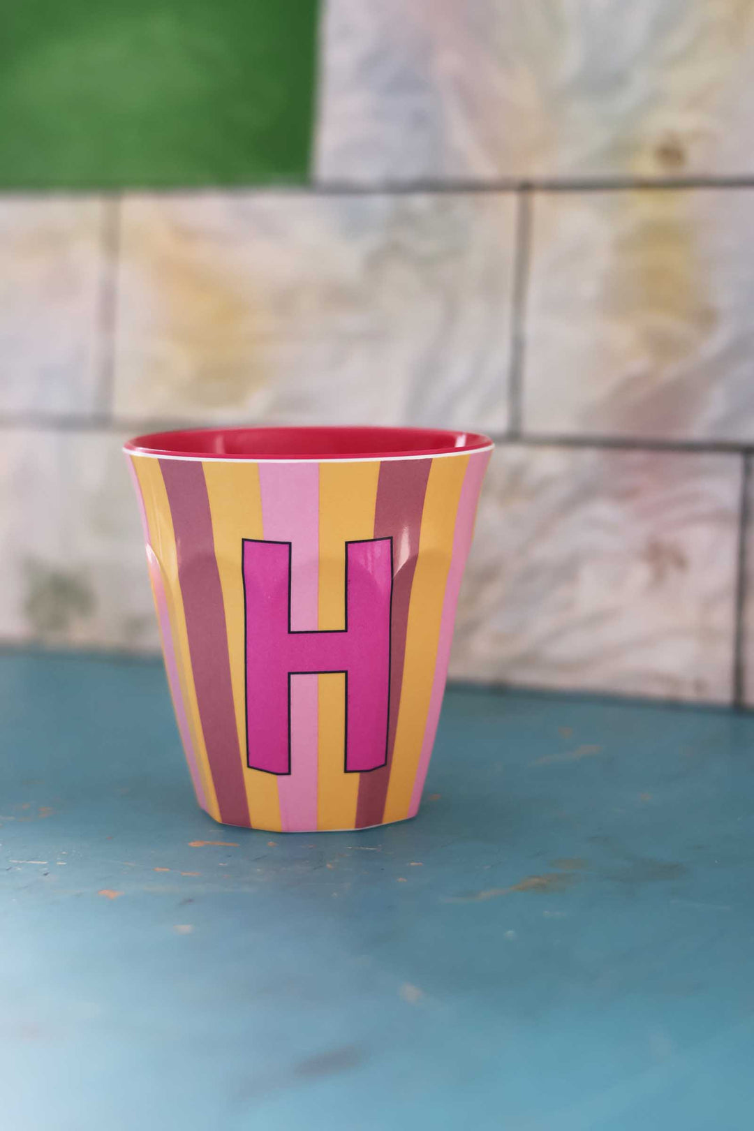 Colorful cup with a pink letter 'H' on a blue surface with a stone wall background