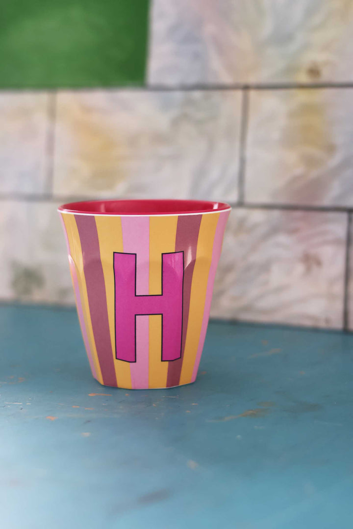 Colorful cup with a pink letter 'H' on a blue surface with a stone wall background