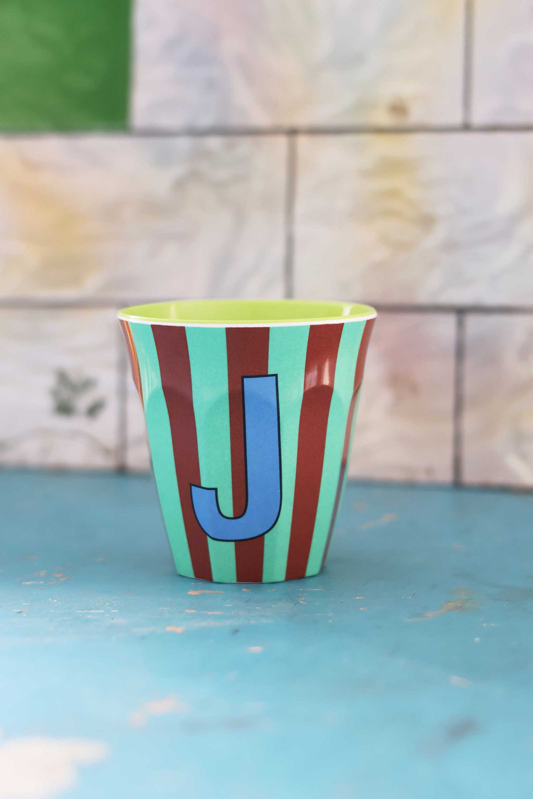 Colorful striped cup with a letter 'J' on a blue surface and tiled wall background