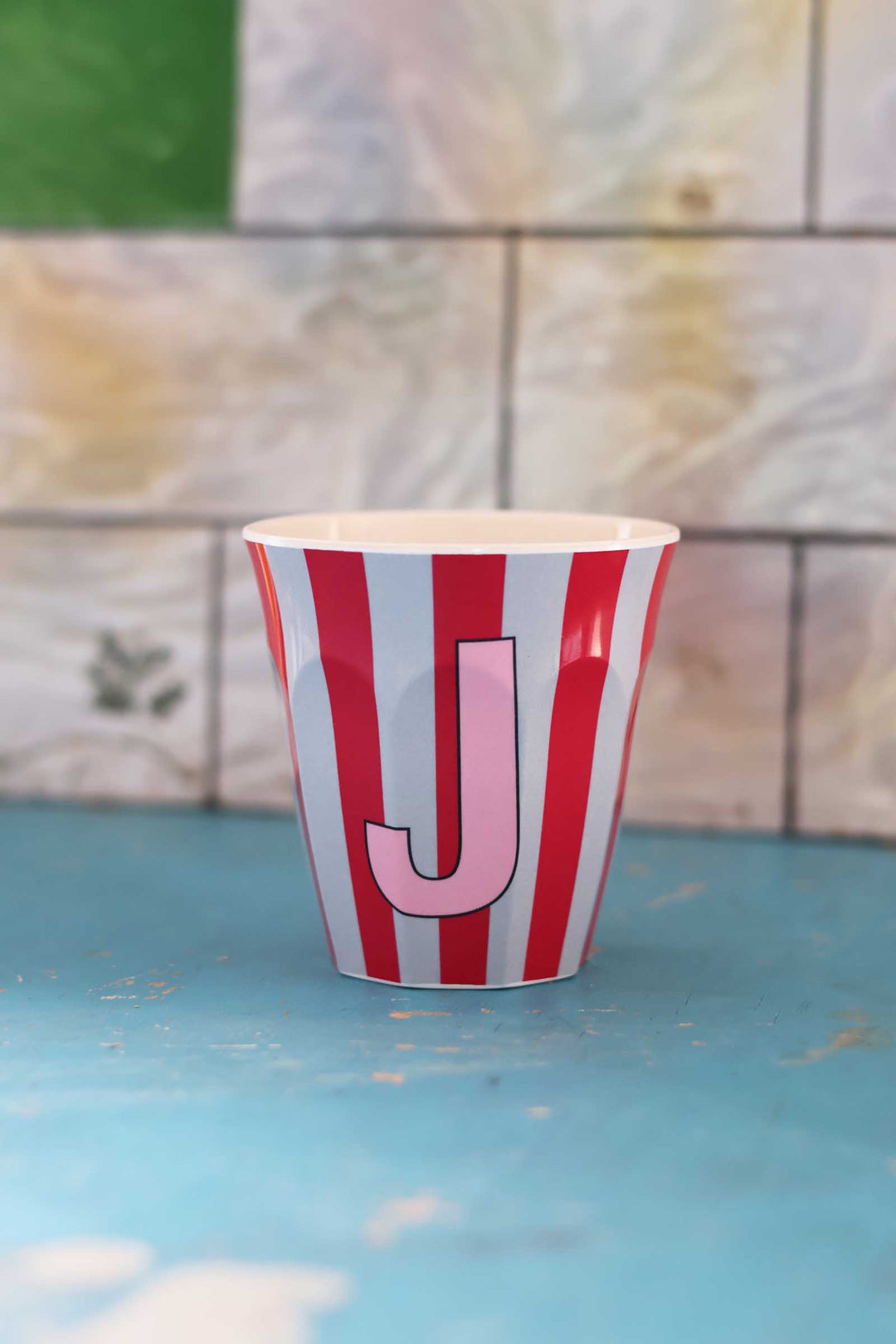 Paper cup with red and white stripes and a pink letter 'J' on a blue surface with a tiled background.