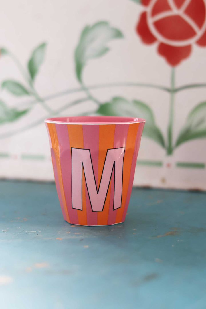 Pink and orange striped cup with letter 'M' on a blue surface, floral patterned background