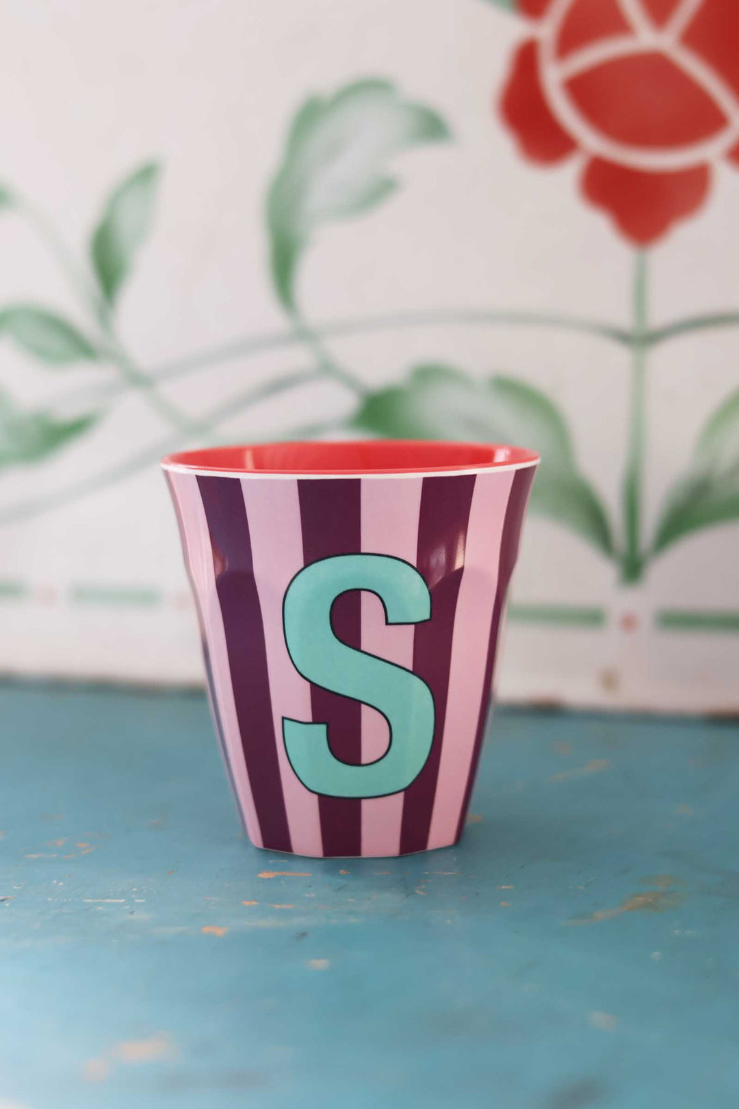 Striped cup with a red interior and turquoise letter 'S' on a blue surface with a floral background.