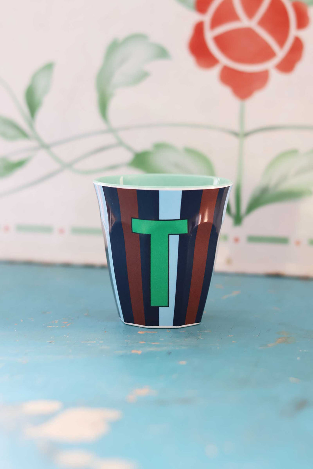 Striped cup with a green letter 'T' on a light blue surface with floral background