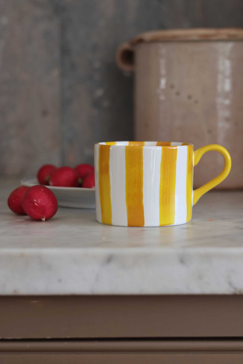 A white ceramic mug with bold yellow vertical stripes and a yellow handle, sitting on a light marble countertop with red radishes and a ceramic jar in the background.